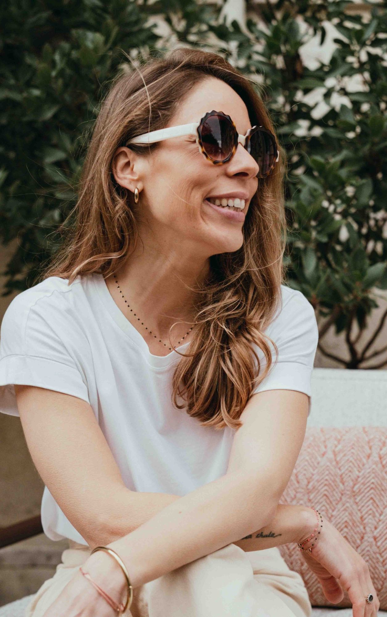 Femme souriante portant des lunettes de soleil, assise sur un canapé avec des plantes en arrière-plan.