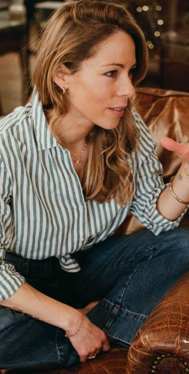 Femme assise sur un canapé, portant une chemise rayée et discutant, cheveux ondulés.